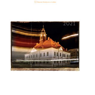 Kalendarz Muzeum Historycznego w Lubinie na 2021 rok. Fotografia nocna przedstawia zabytkowy lubiński ratusz z charakterystyczną wieżą zegarową. Długi czas naświetlania tworzy na zdjęciu świetlne smugi przejeżdżających pojazdów. Nad fotografią umieszczono logo Muzeum Historycznego w Lubinie oraz napis „2021”.